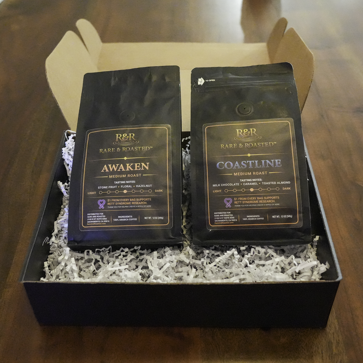 Two black coffee bags from Rare & Roasted, labeled Awaken and Coastline, displayed in a black gift box filled with white crinkle-cut paper on a wooden table.