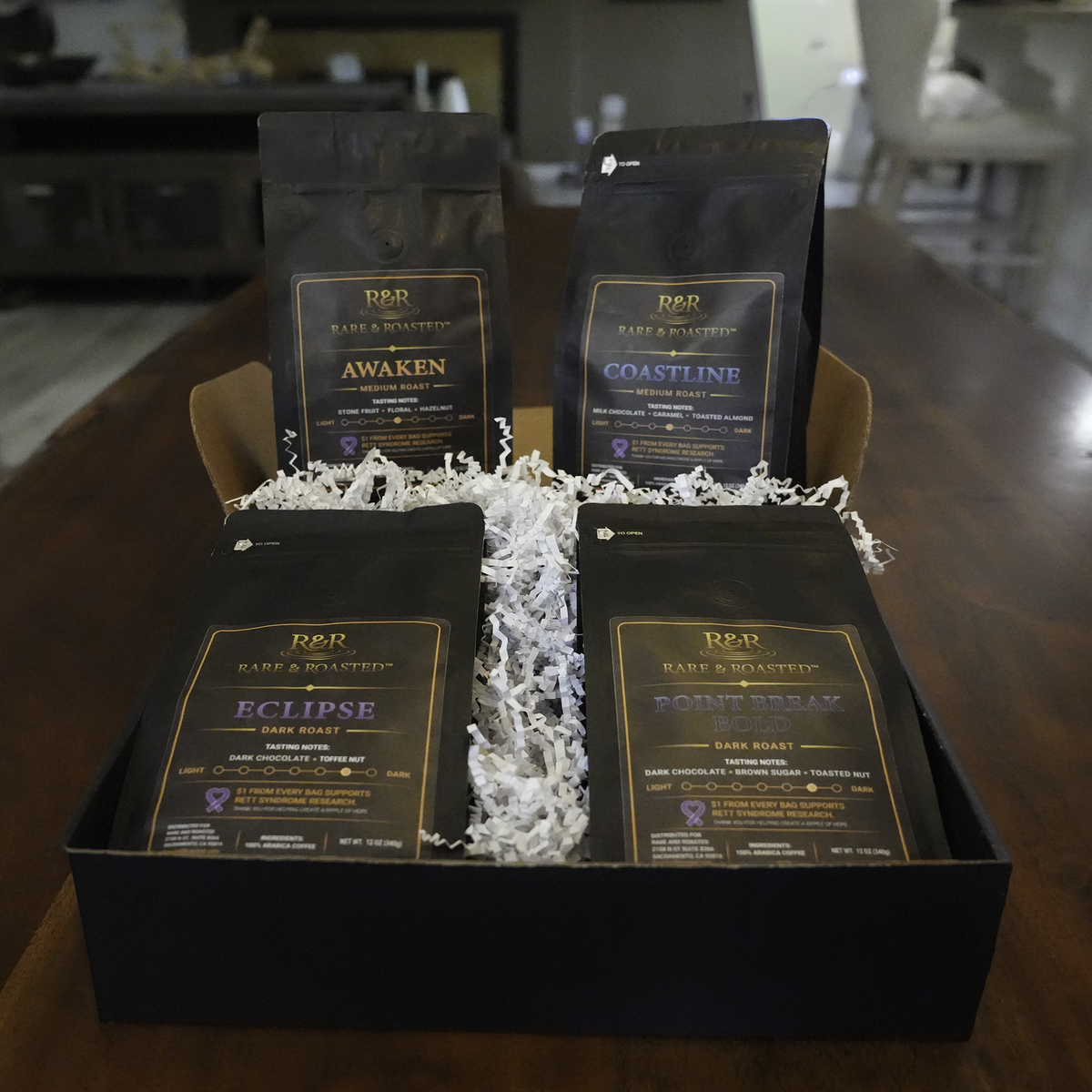 A black gift box containing four bags of R&R Coffee Roasters dark roast coffee in different flavor profiles: Awaken, Coastline, Eclipse, and Point Break Bold, arranged on white shredded crinkle-cut paper on a wooden table.