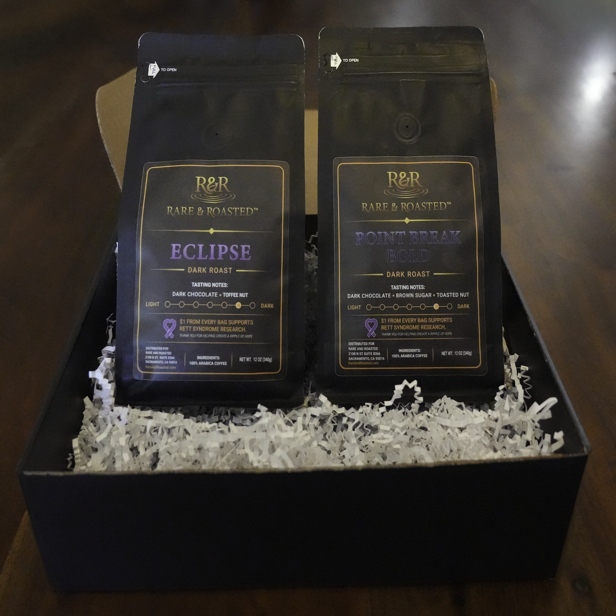 Two bags of Rare & Roasted coffee in a black gift box with white crinkle paper on a dark wooden surface. The bags are labeled Eclipse Dark Roast and Point Break Bold Dark Roast.